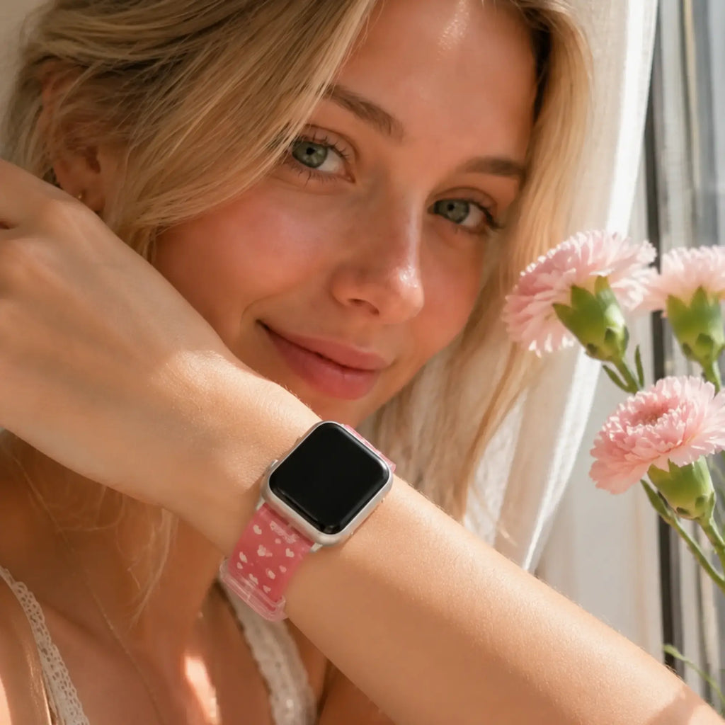 Close-up of the Amour pink heart Apple Watch band worn beside pink flowers.