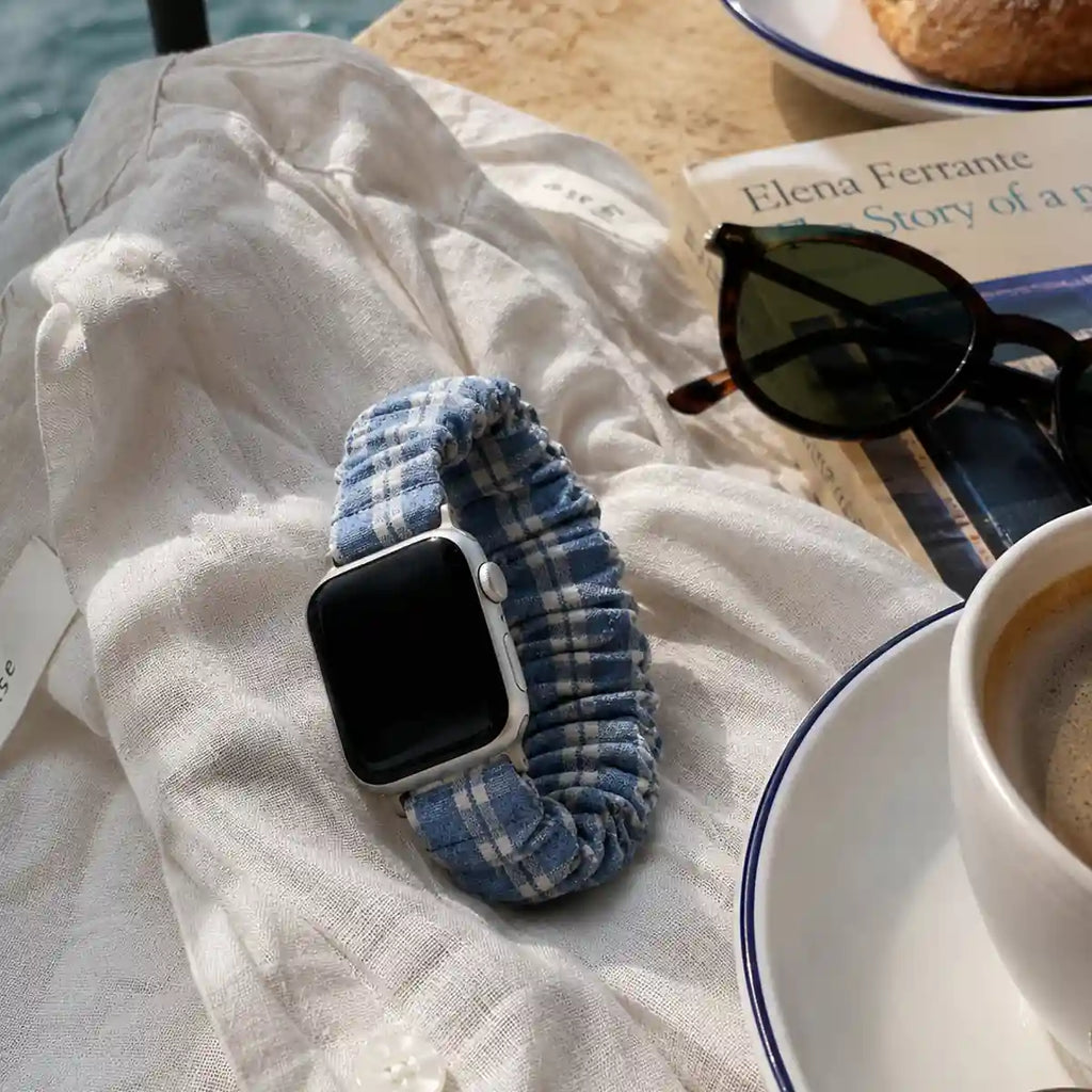 Camille blue gingham scrunchie Apple Watch band in a seaside café flat lay with coffee and sunglasses.
