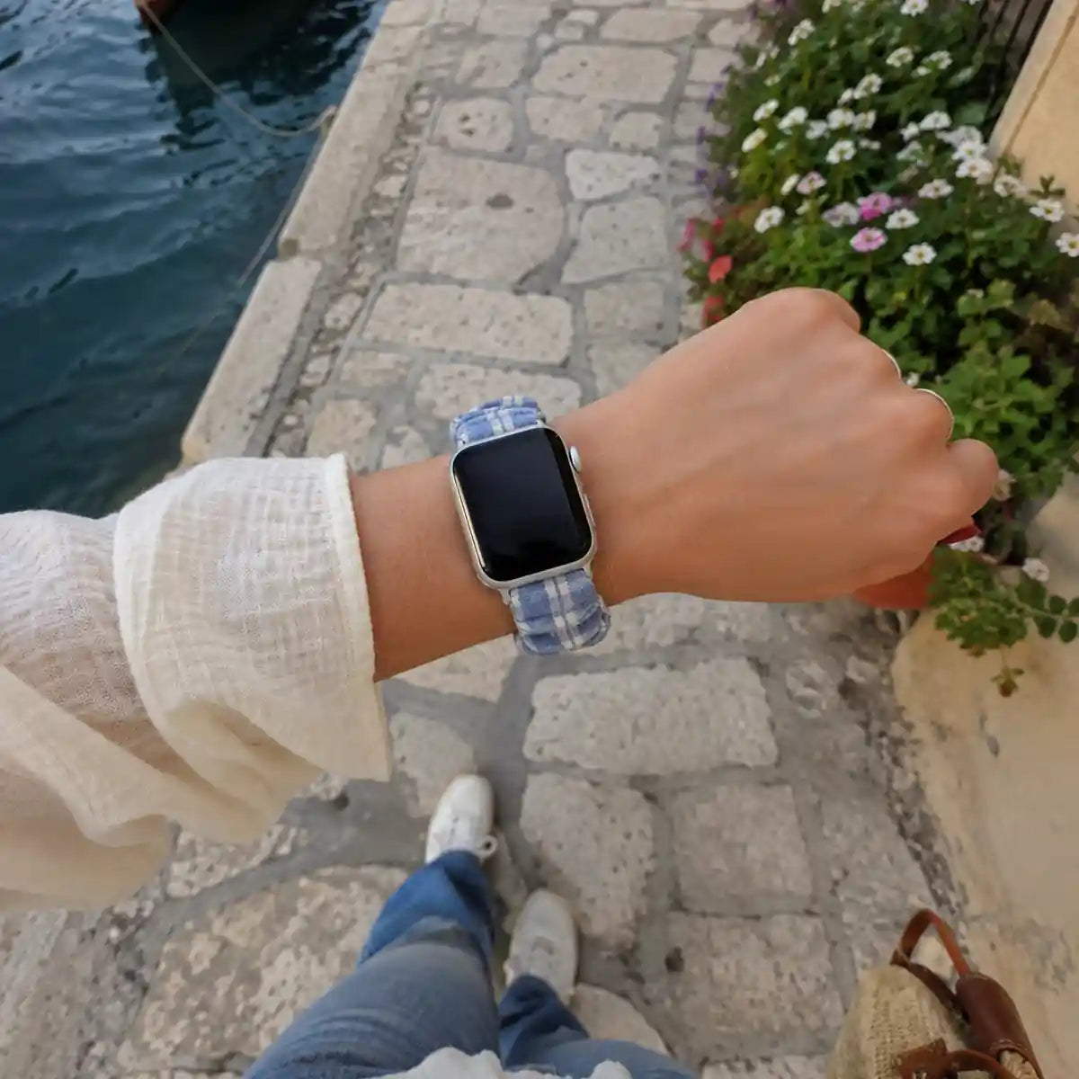 Camille blue gingham scrunchie Apple Watch band shown on the wrist beside a flower-lined harbor.