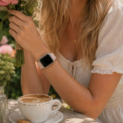 Close-up of the Luna blush silicone Apple Watch band worn beside flowers and coffee.