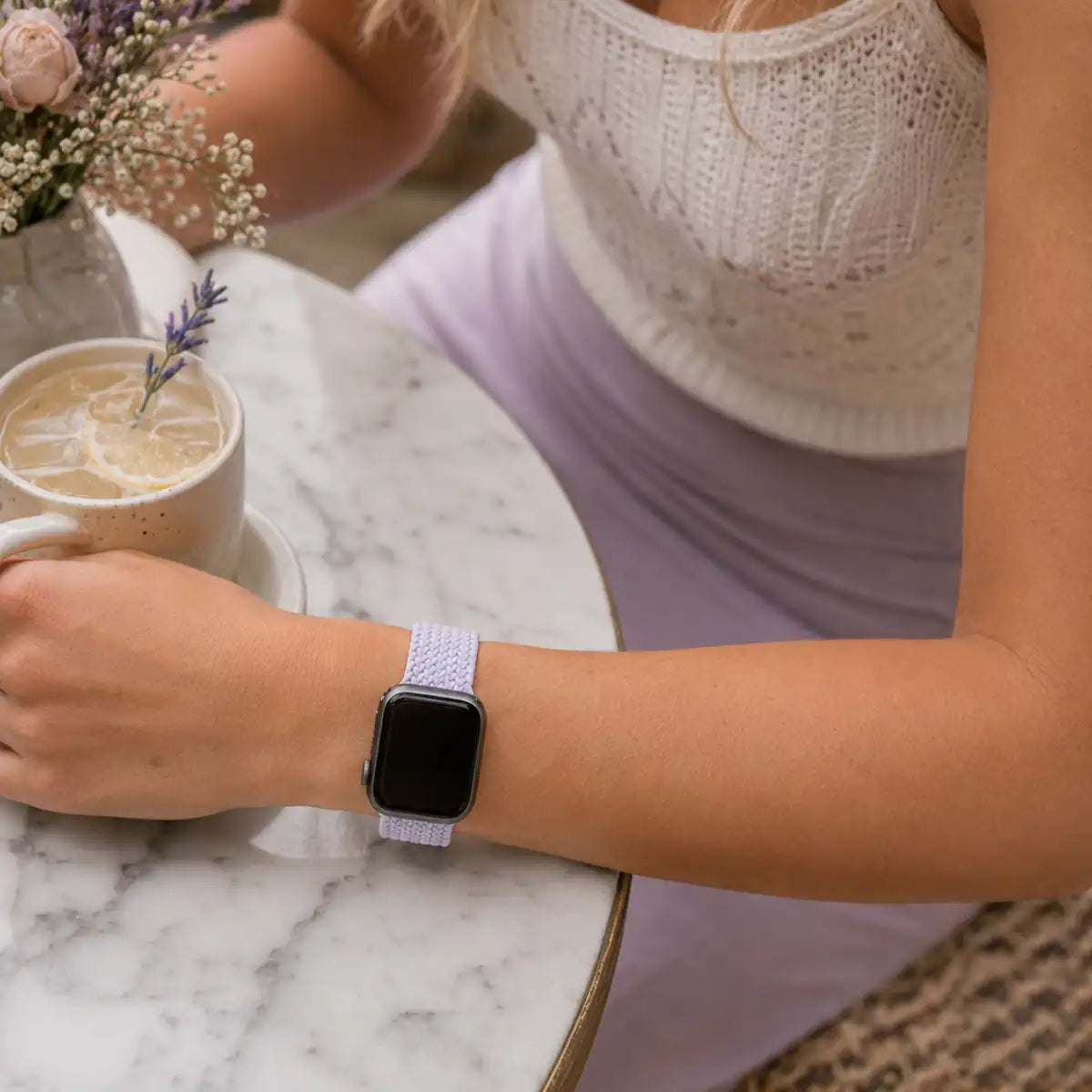 Close-up of the Soline lavender braided loop Apple Watch band worn at a marble café table.